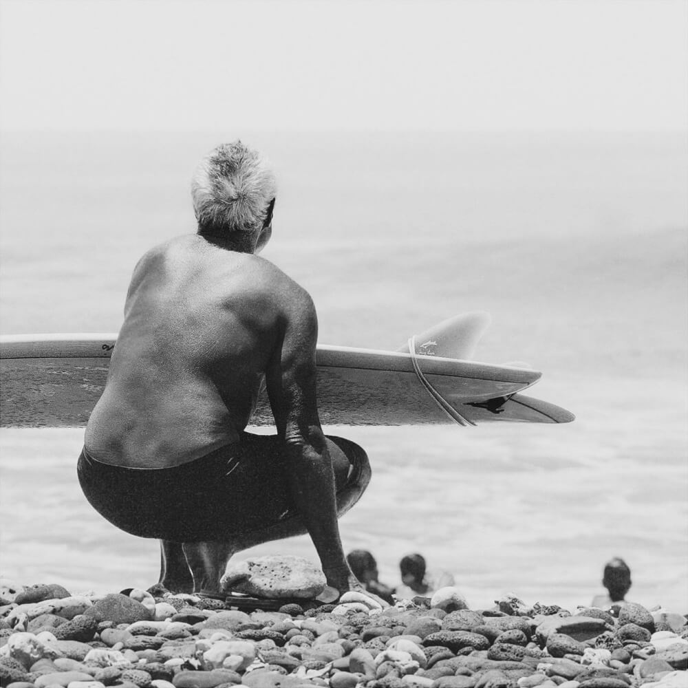 Old man surfer crouching on beach with surfboard